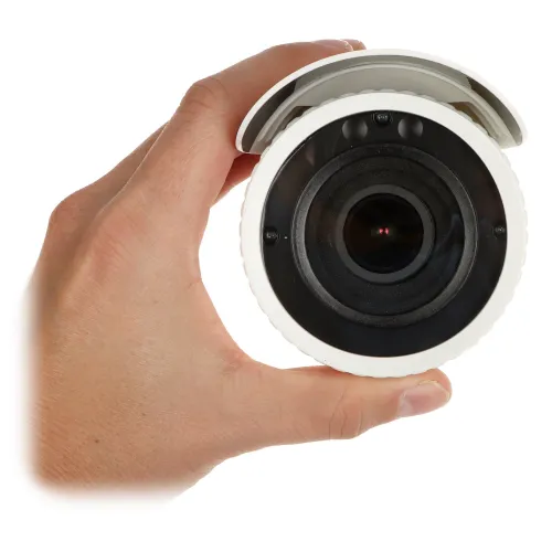 Osoba trzyma w dłoni białą kamerę monitoringu z widocznym obiektywem skierowanym w stronę aparatu. Kamera ma cylindryczny kształt i osłonę przeciwsłoneczną.