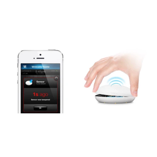 Na zdjęciu znajduje się czujnik zalania wodą Fibaro, który jest biały i ma okrągły kształt. Obok niego widoczny jest smartfon z aplikacją pokazującą alert o wykryciu zalania.