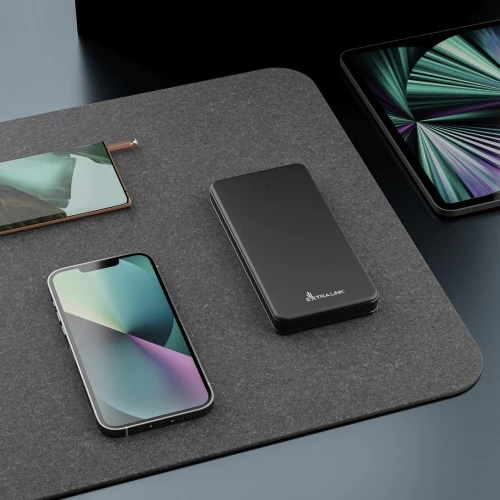 Na zdjęciu znajduje się czarny powerbank marki Extralink umieszczony na szarej powierzchni obok smartfona i tabletu. Tło jest minimalistyczne, co podkreśla elegancki design urządzeń elektronicznych.