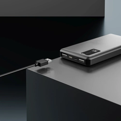 Na zdjęciu znajduje się czarny powerbank z wyświetlaczem cyfrowym pokazującym poziom naładowania, umieszczony na ciemnej, geometrycznej powierzchni. Obok powerbanka leży kabel USB, gotowy do podłączenia.