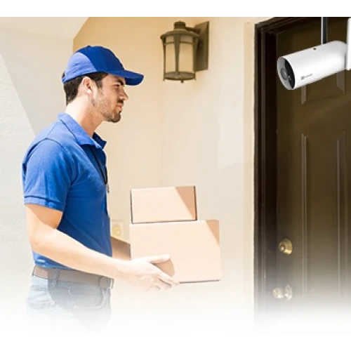 Mężczyzna w niebieskiej czapce i koszulce dostarcza paczki pod drzwi domu. Nad drzwiami zamontowana jest biała kamera monitoringu.