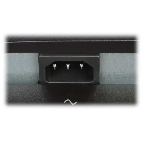 Zdjęcie przedstawia zbliżenie na gniazdo zasilania z trzema metalowymi bolcami, umieszczone w czarnej obudowie. Tło jest rozmyte, a na dolnej części obudowy widoczny jest symbol fali oznaczający prąd zmienny.