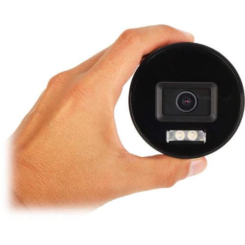 Zbliżenie na kamerę IP trzymaną w dłoni, z widocznym obiektywem i dwoma diodami LED poniżej. Kamera ma czarną, okrągłą obudowę.