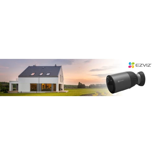 Na zdjęciu znajduje się czarna kamera WiFi marki EZVIZ o rozdzielczości 4MP, zasilana bateriami. W tle widoczny jest nowoczesny dom jednorodzinny na tle zielonego krajobrazu.