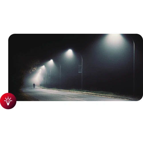 Zdjęcie przedstawia nocną ulicę oświetloną szeregiem latarni, z pojedynczą postacią idącą w oddali. Atmosfera jest tajemnicza, z mgłą unoszącą się wokół latarni.